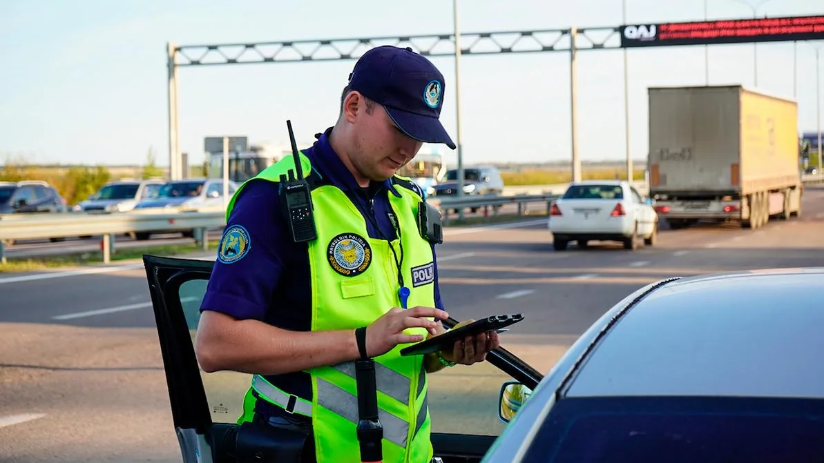 Сотрудник дорожной полиции останавливает автомобиль на трассе, рядом видно дорожный знак ограничения скорости и пейзаж Казахстана.