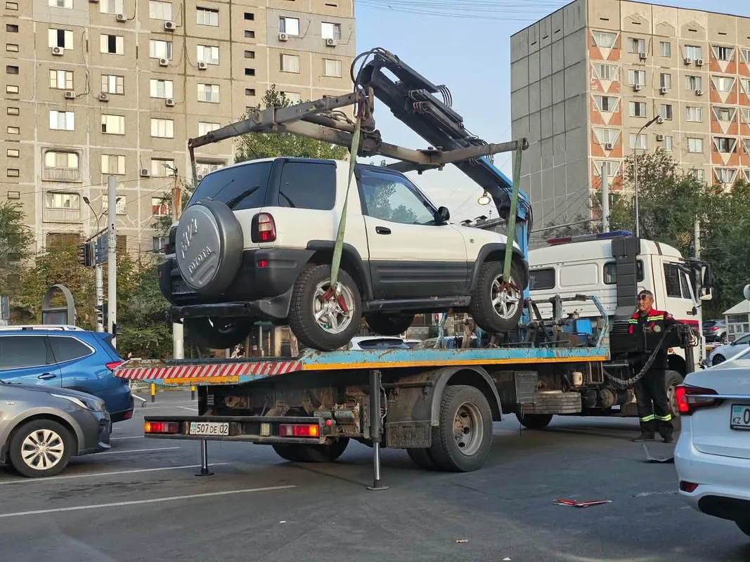 Эвакуатор перевозит легковой автомобиль по городской улице в Алматы, на заднем плане видны жилые дома и другие машины