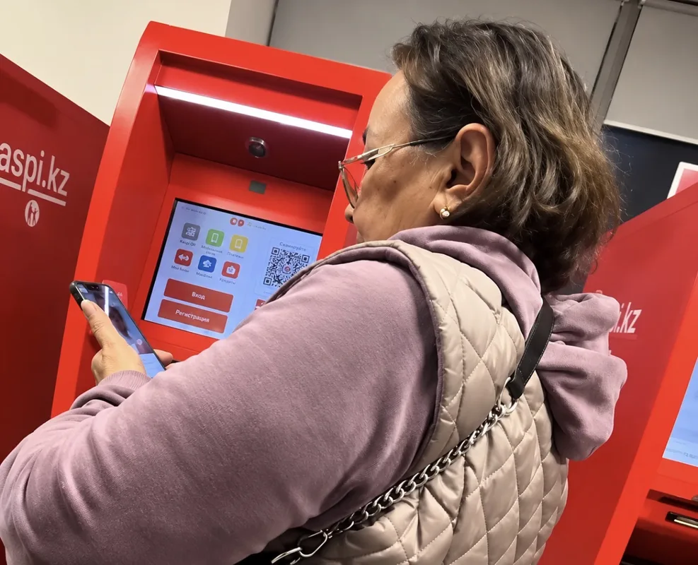 Пожилая женщина у банкомата Kaspi, общается по видеосвязи, рядом стоят люди